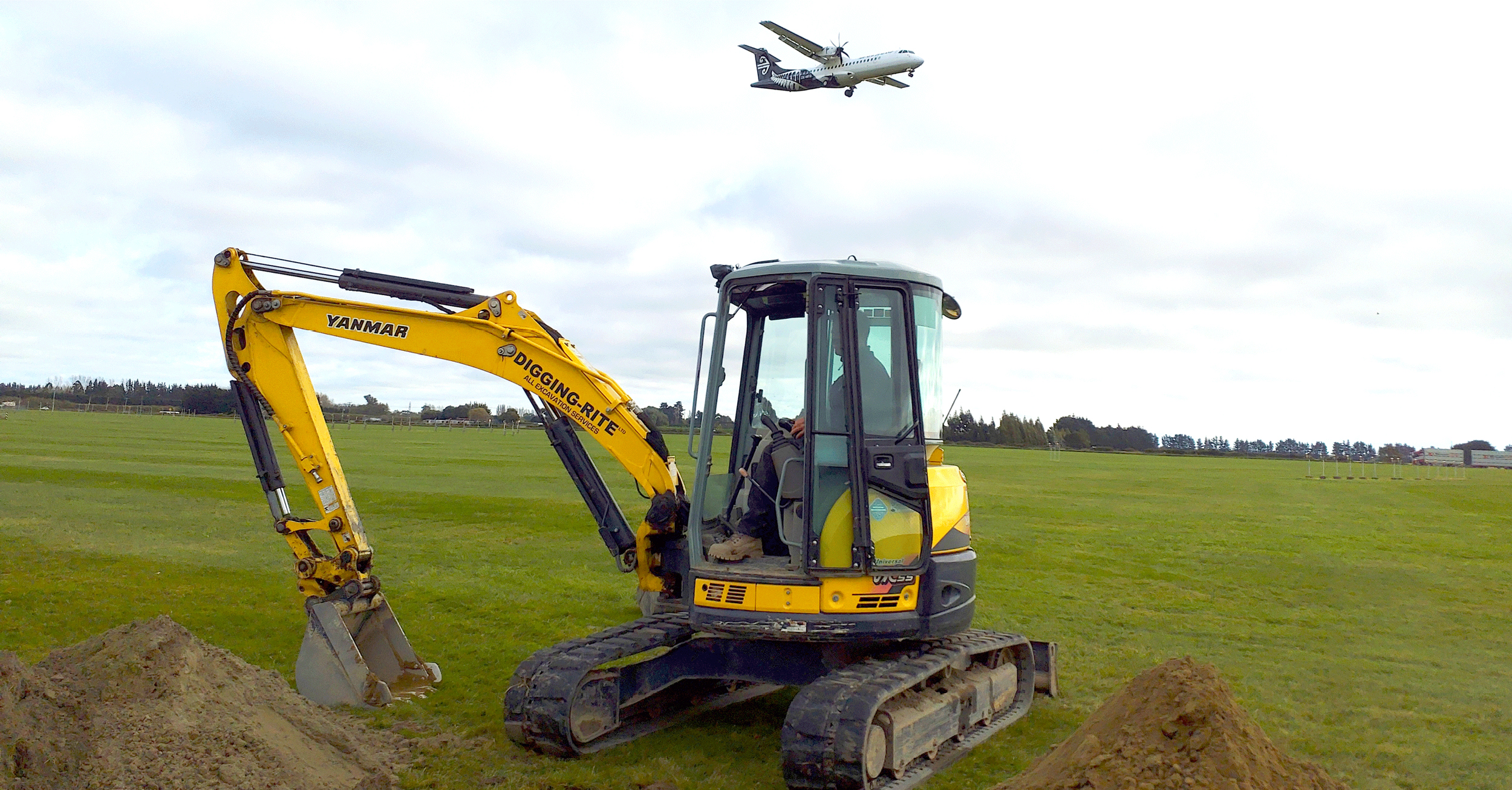 Digger breaking earth at Kōwhai Park Solar Farm in Ōtautahi Christchurch, with aeroplane flying overhead