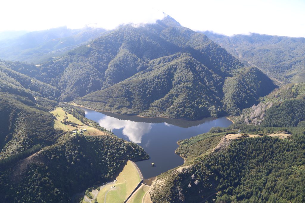 Aerial view of the Maitai Dam and its reservoir surrounded by forested hills and mountains in New Zealand.
